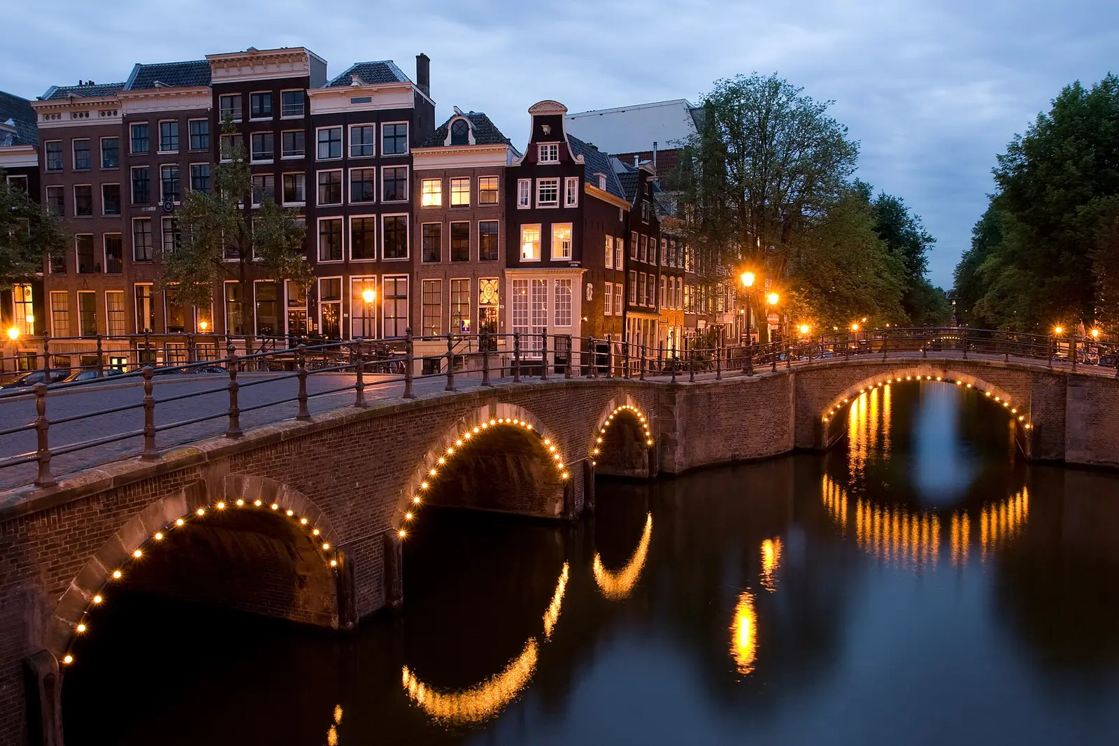 Arched brick bridges lit up at dusk where two canals meet, with gabled canal houses behind