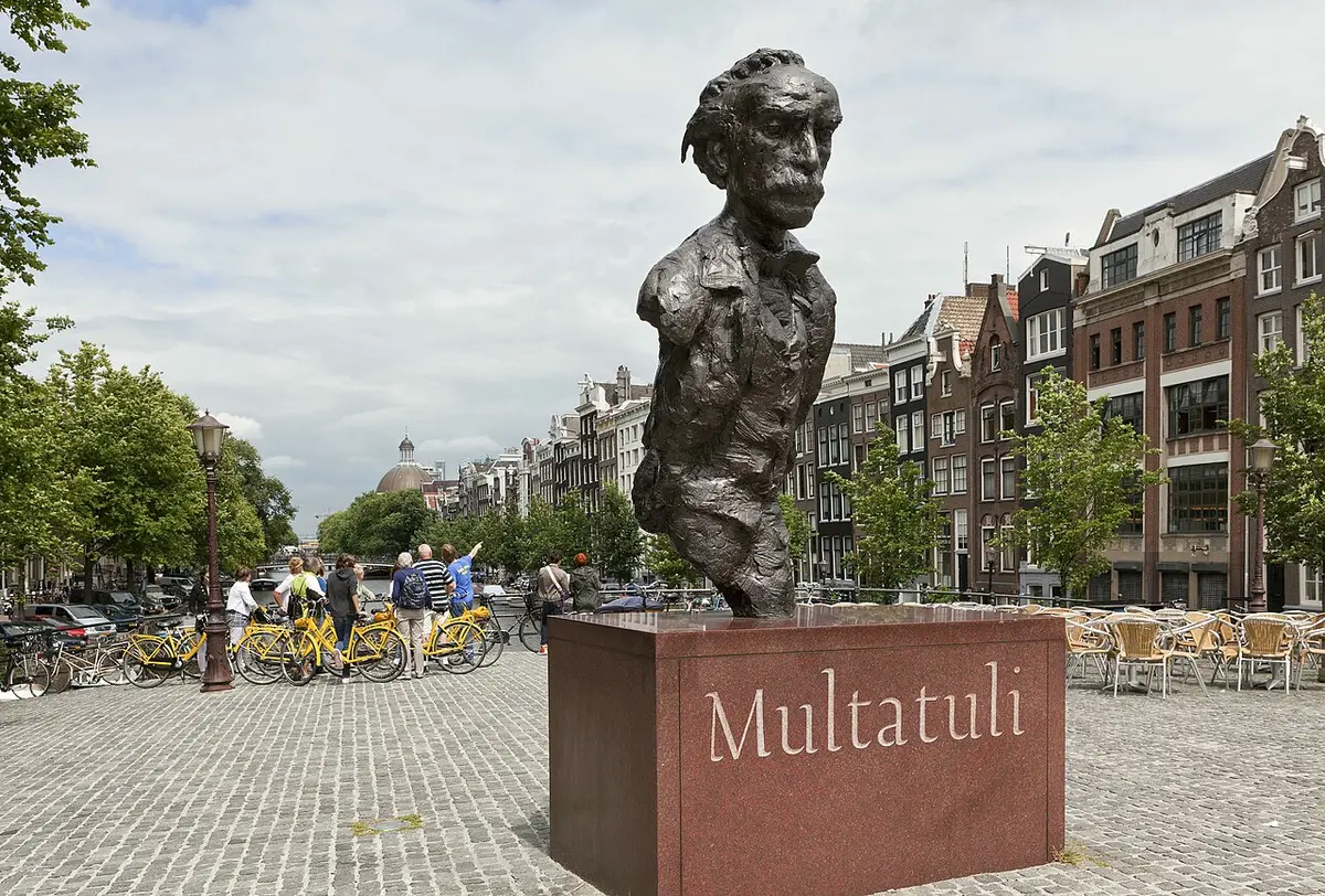 A bronze bust of Multatuli on a red stone plinth on the Torensluis bridge, canal houses behind