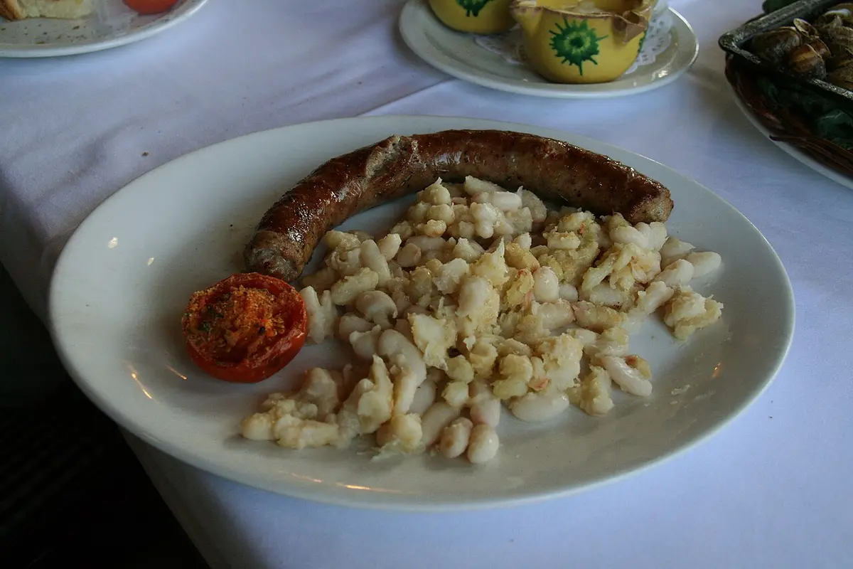 A grilled botifarra sausage with white beans and half a tomato on a plate