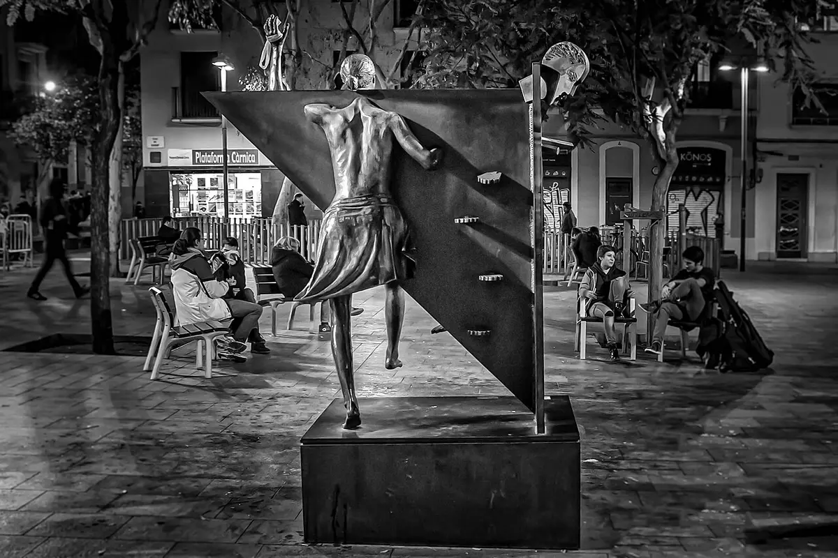 Black and white photograph of a bronze woman breaking out of a steel triangle in a paved square at night
