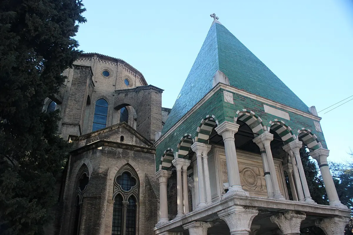 A medieval tomb raised on columns under a green-tiled pyramid roof, beside the brick apse of San Francesco