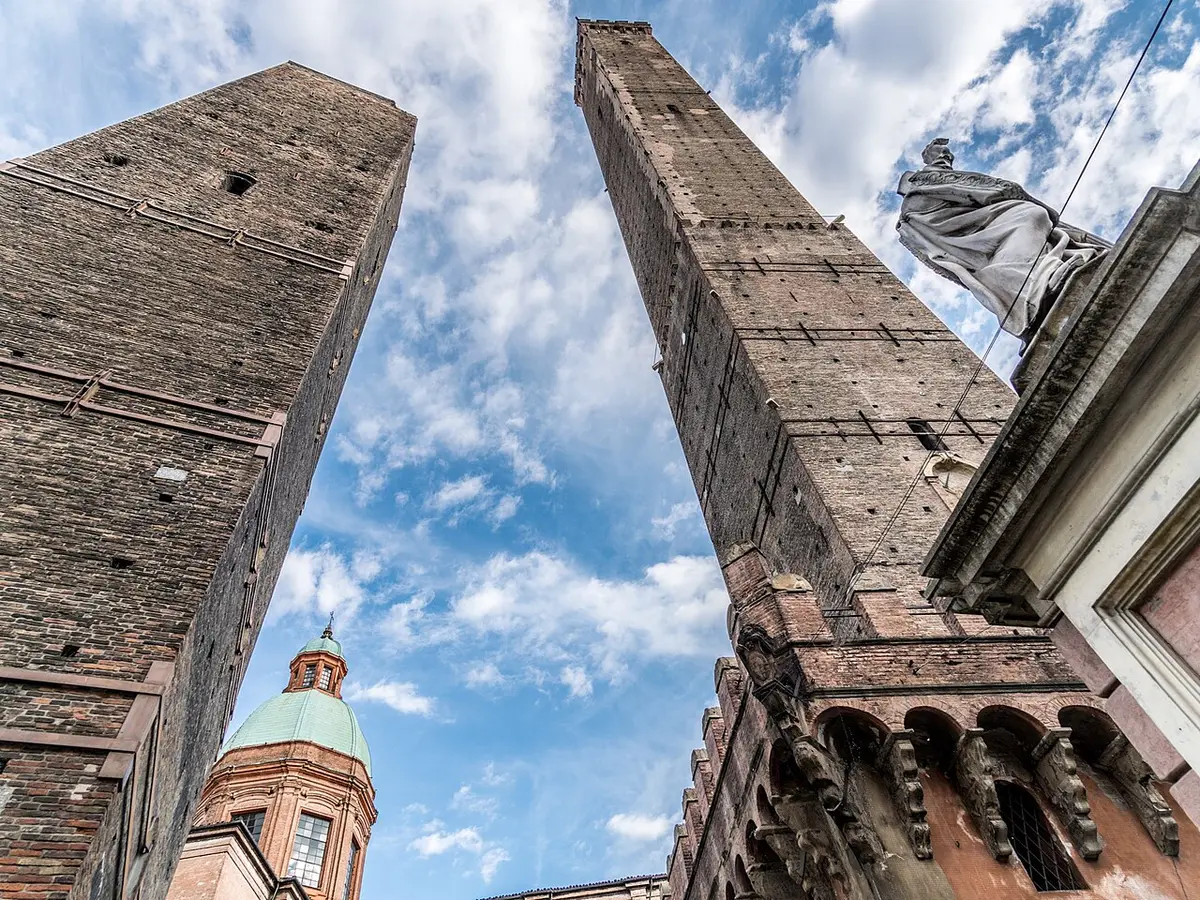 The Asinelli and Garisenda towers seen from below against a blue sky, a statue of San Petronio beside them