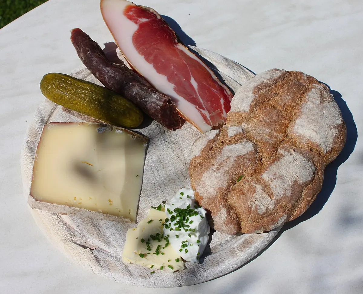 A wooden board with speck, a smoked Kaminwurz sausage, a gherkin, cheese, a rye loaf, butter and quark with chives