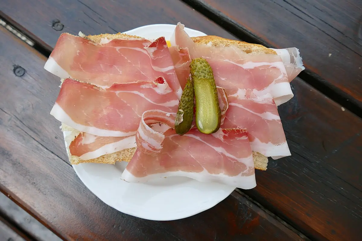 Thin slices of speck on bread with two small gherkins, on a white plate