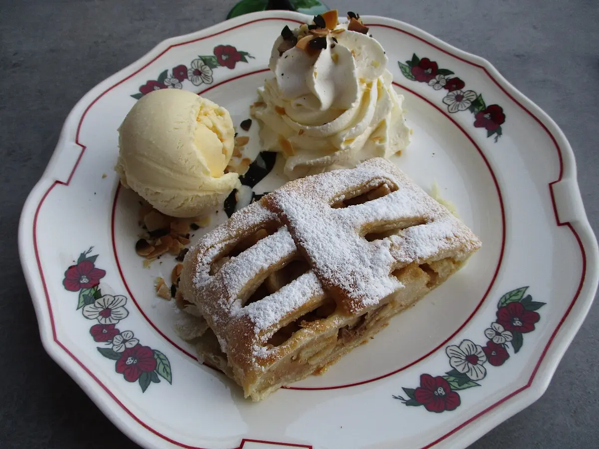 A slice of apple strudel with a lattice top, dusted with sugar, next to vanilla ice cream and whipped cream