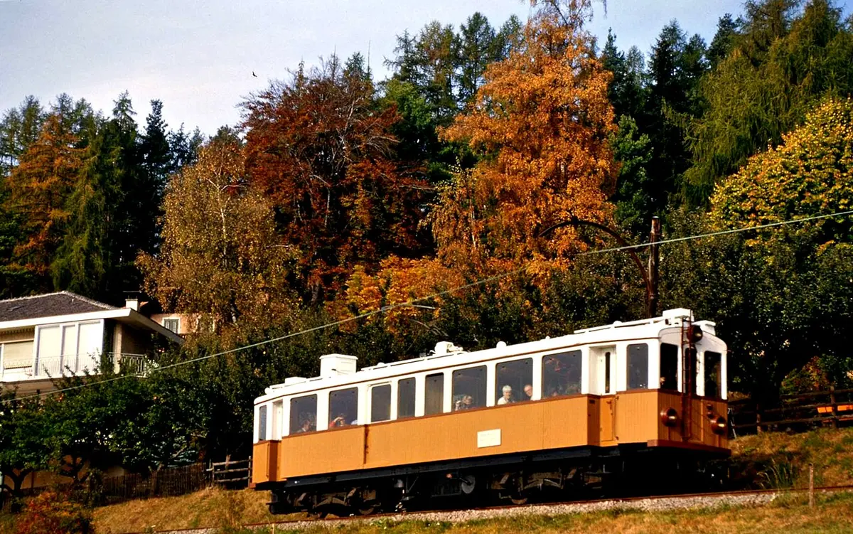 An orange and white historic railcar of the Renon railway among autumn larches