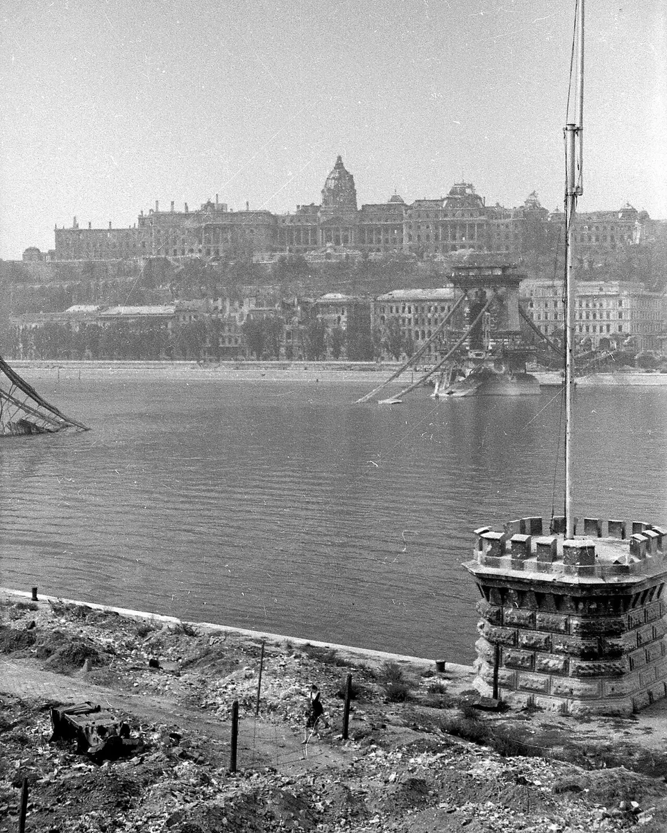 Black and white photograph of the Chain Bridge after it was blown up: a stone tower standing in the river, the chains in the water, the ruined palace on the hill behind