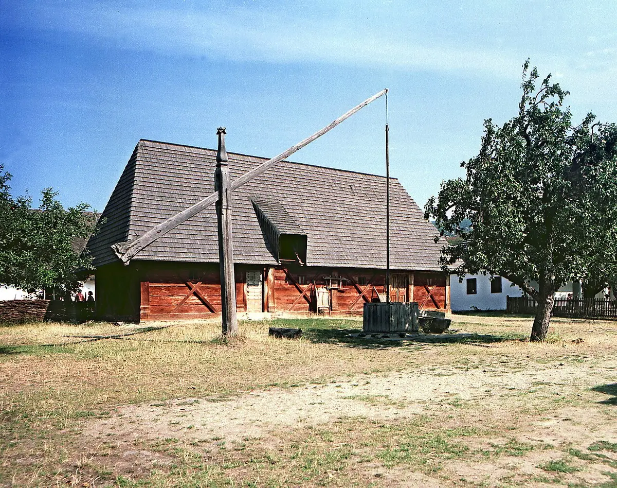 An old wooden farmhouse with a shingle roof and a well sweep in front, at the open-air museum