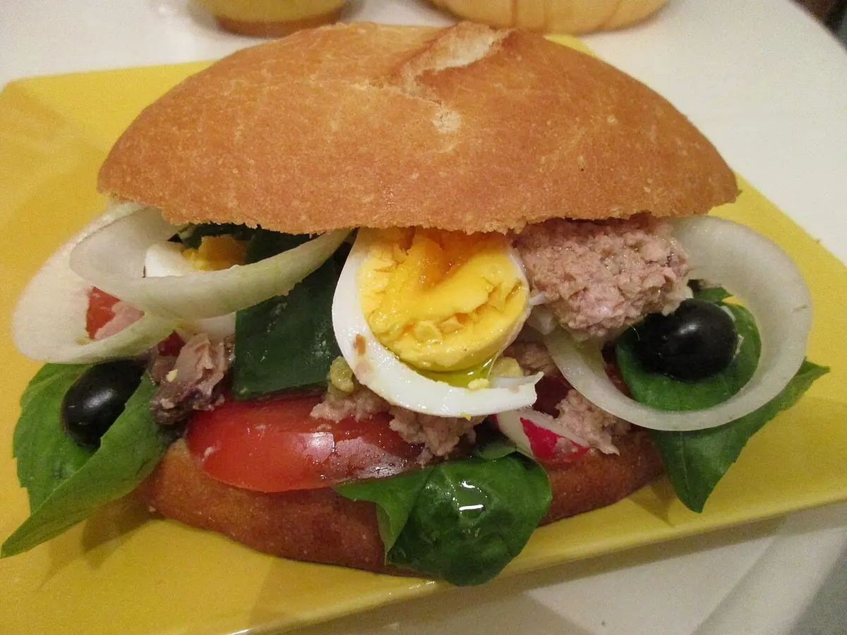 A round bread roll filled with tuna, hard-boiled egg, tomato, onion, black olives and leaves