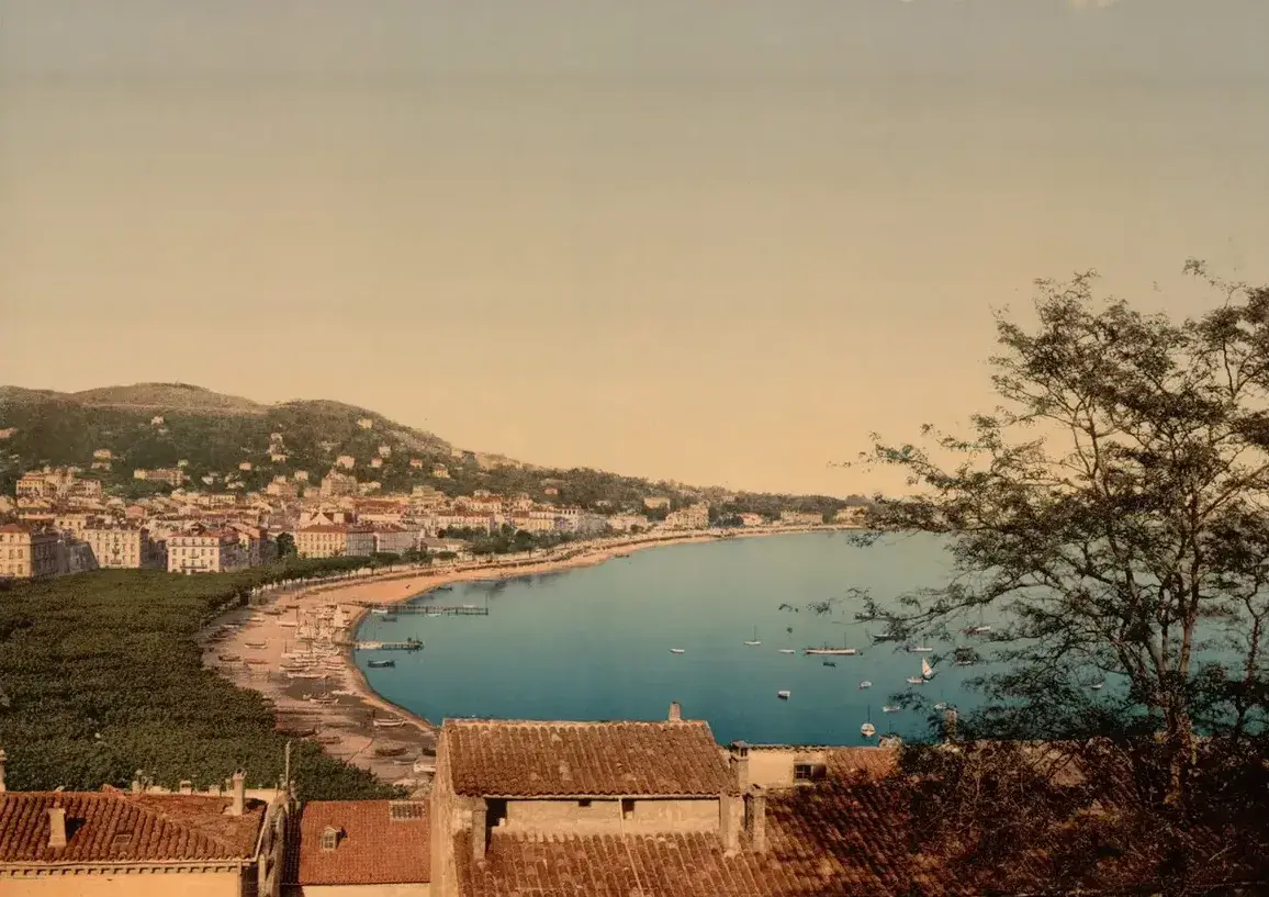 Hand-colored view over old roofs to the curve of the bay of Cannes, its beach and villas under wooded hills