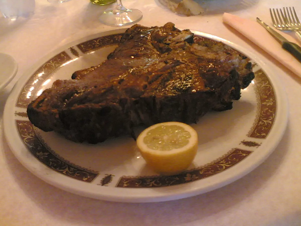 A thick grilled T-bone steak on a white plate with a wedge of lemon