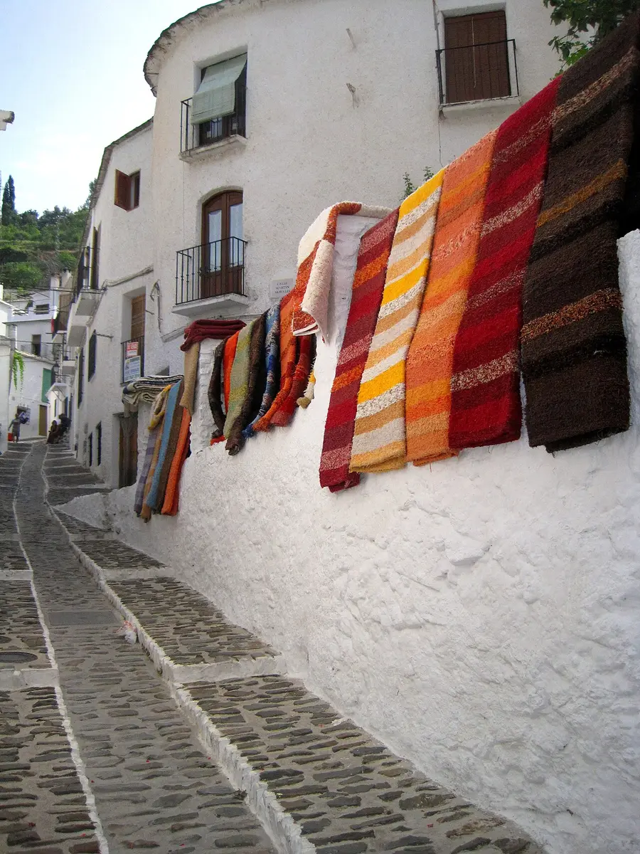 Striped woven rugs in red, orange and brown hung on a white wall along a stepped cobbled street
