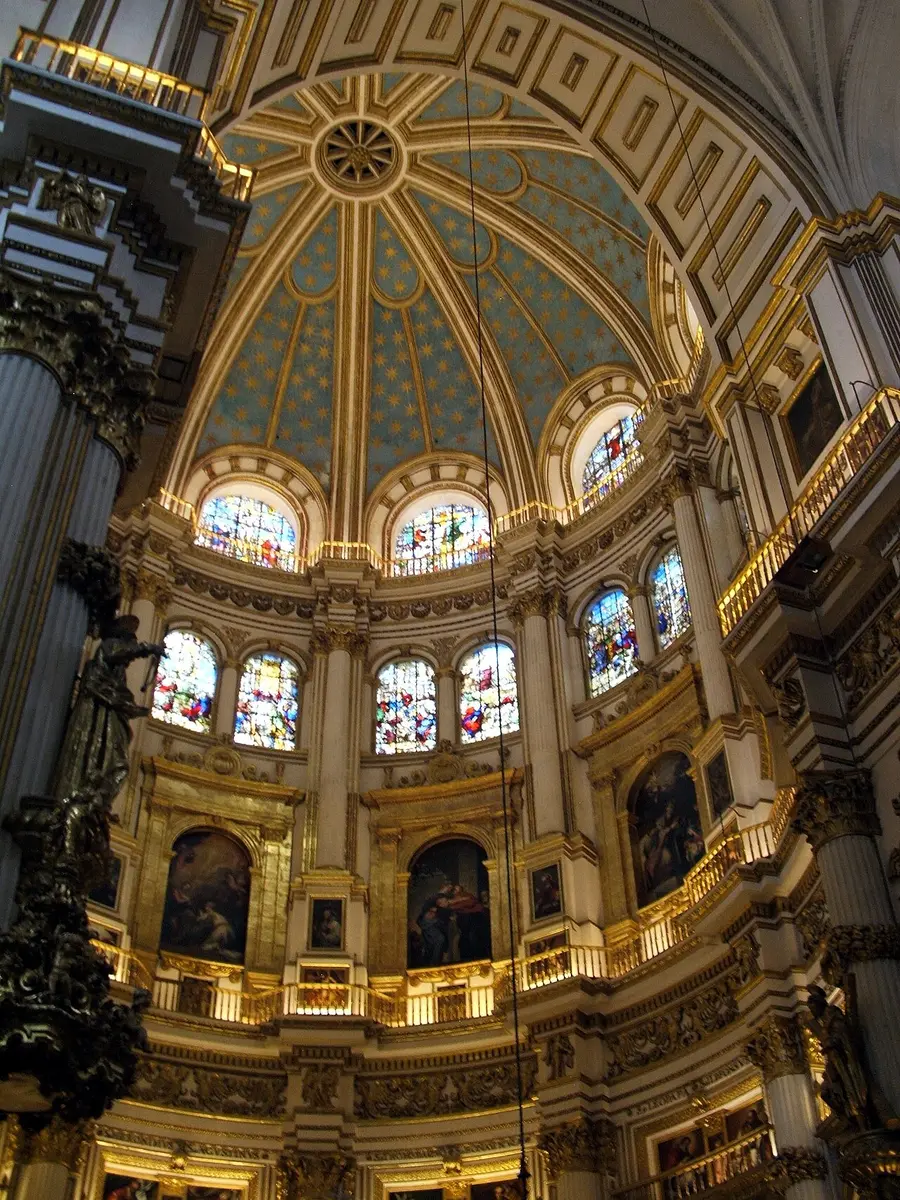 The white and gold main chapel of Granada Cathedral, with stained glass windows and large paintings in tiers