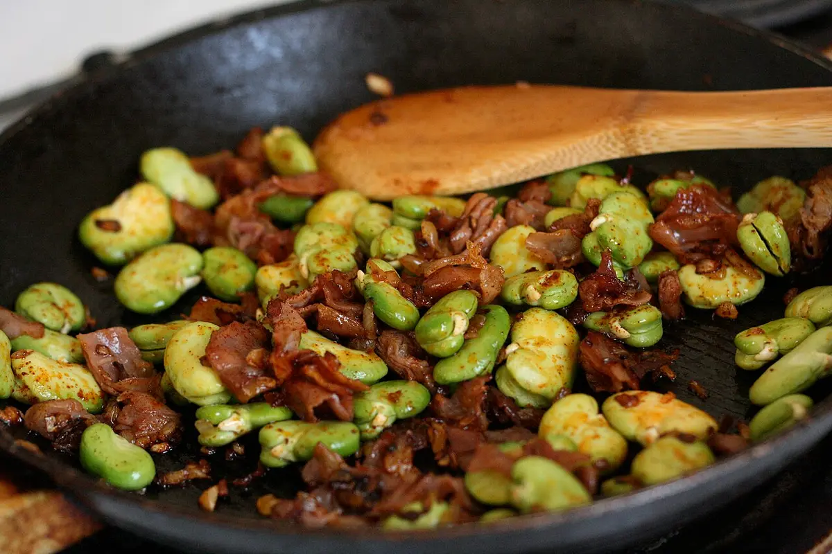 Broad beans fried with pieces of cured ham in a black pan, a wooden spoon resting on top
