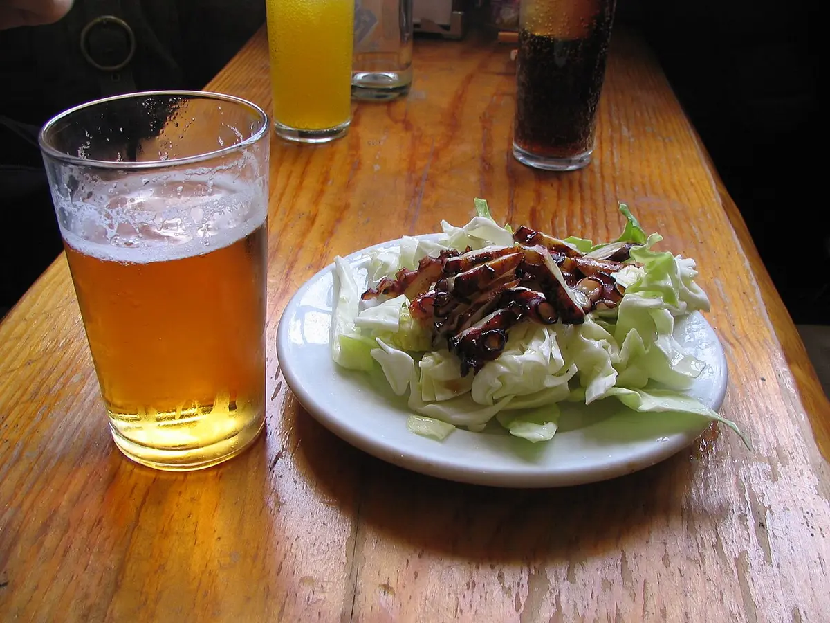 A glass of beer beside a plate of dried octopus on lettuce on a wooden bar