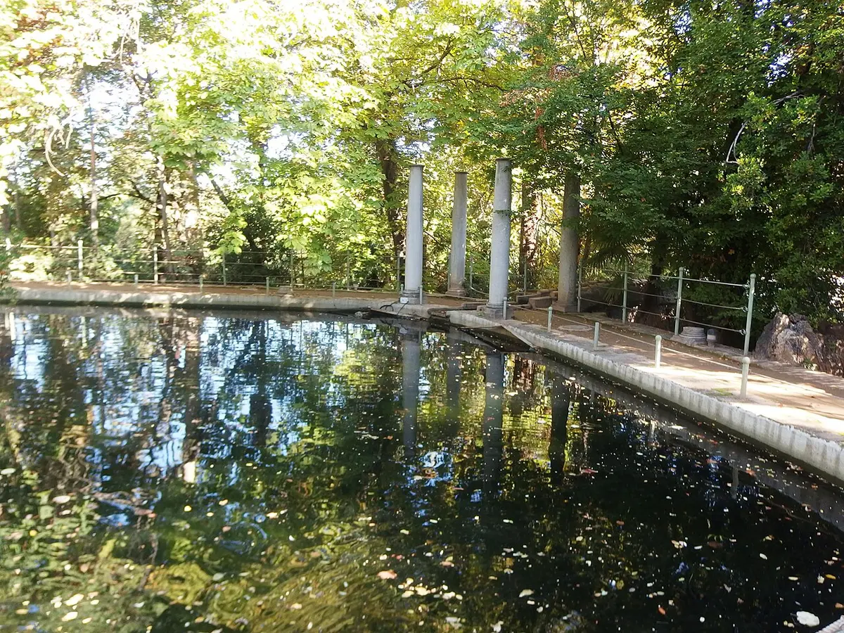A still pond reflecting trees and a row of stone columns in the gardens of the Carmen de los Mártires