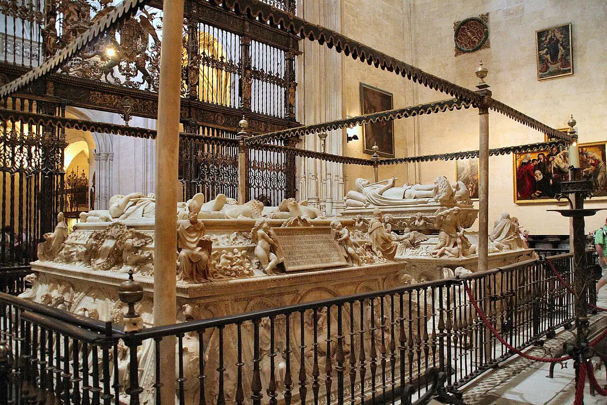 White marble tombs with carved effigies behind a low railing, a gilded iron screen beyond them