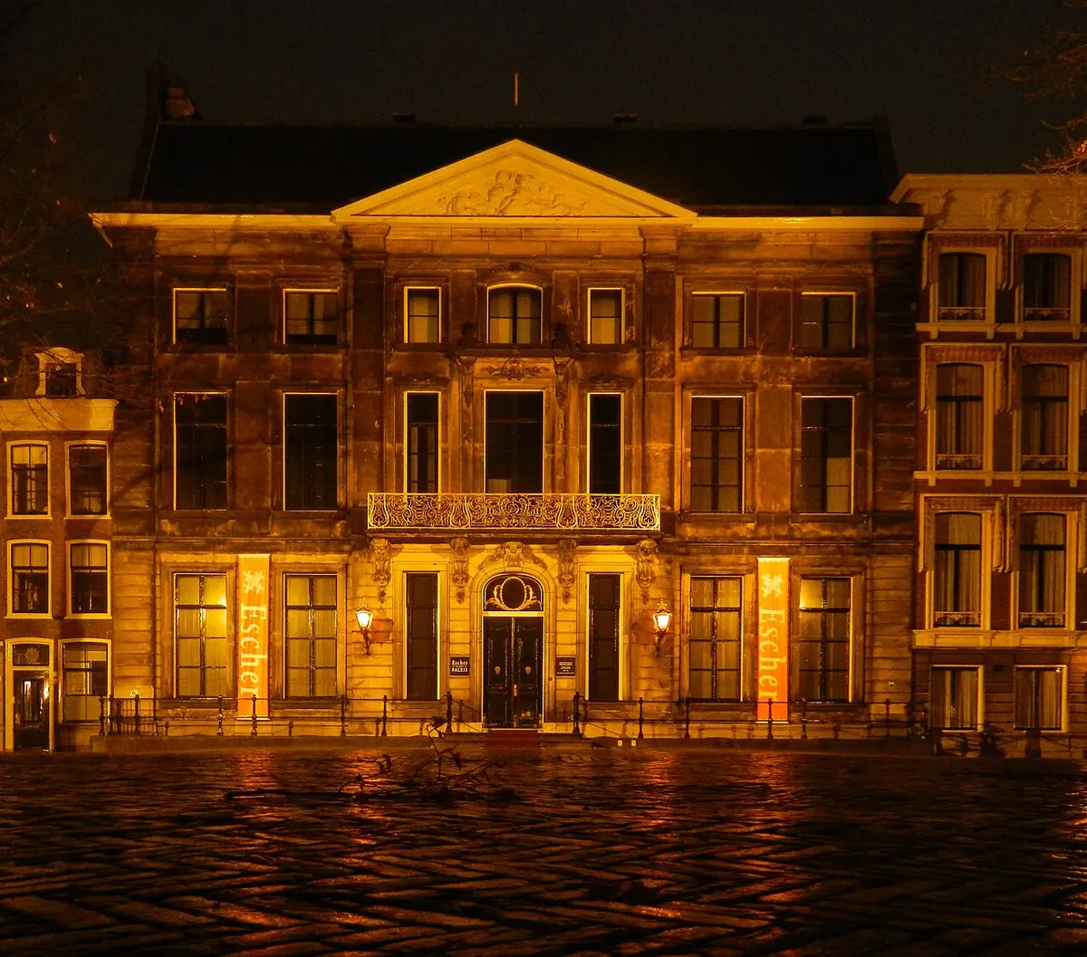 The Lange Voorhout Palace lit up at night, with two tall Escher banners by the door