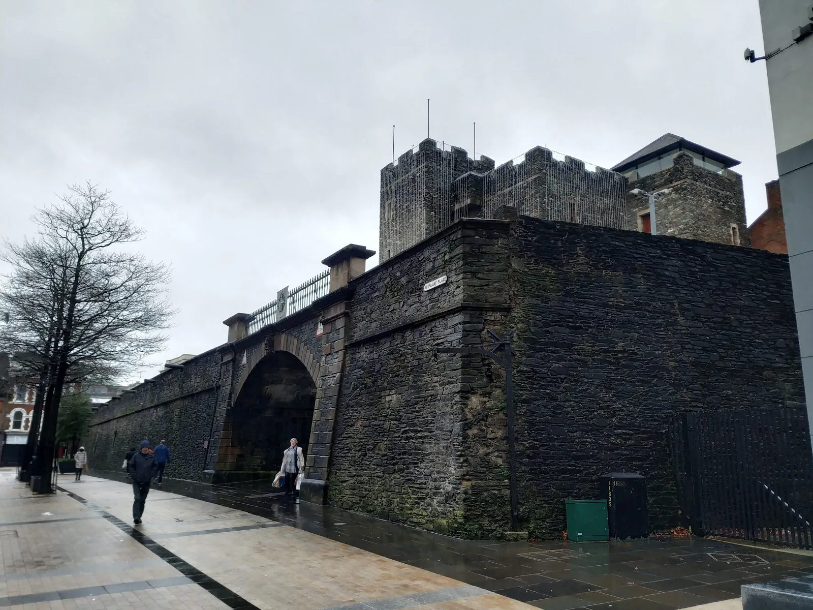 The dark stone city walls of Derry with a gate arch, a lit pavement in front on a rainy day
