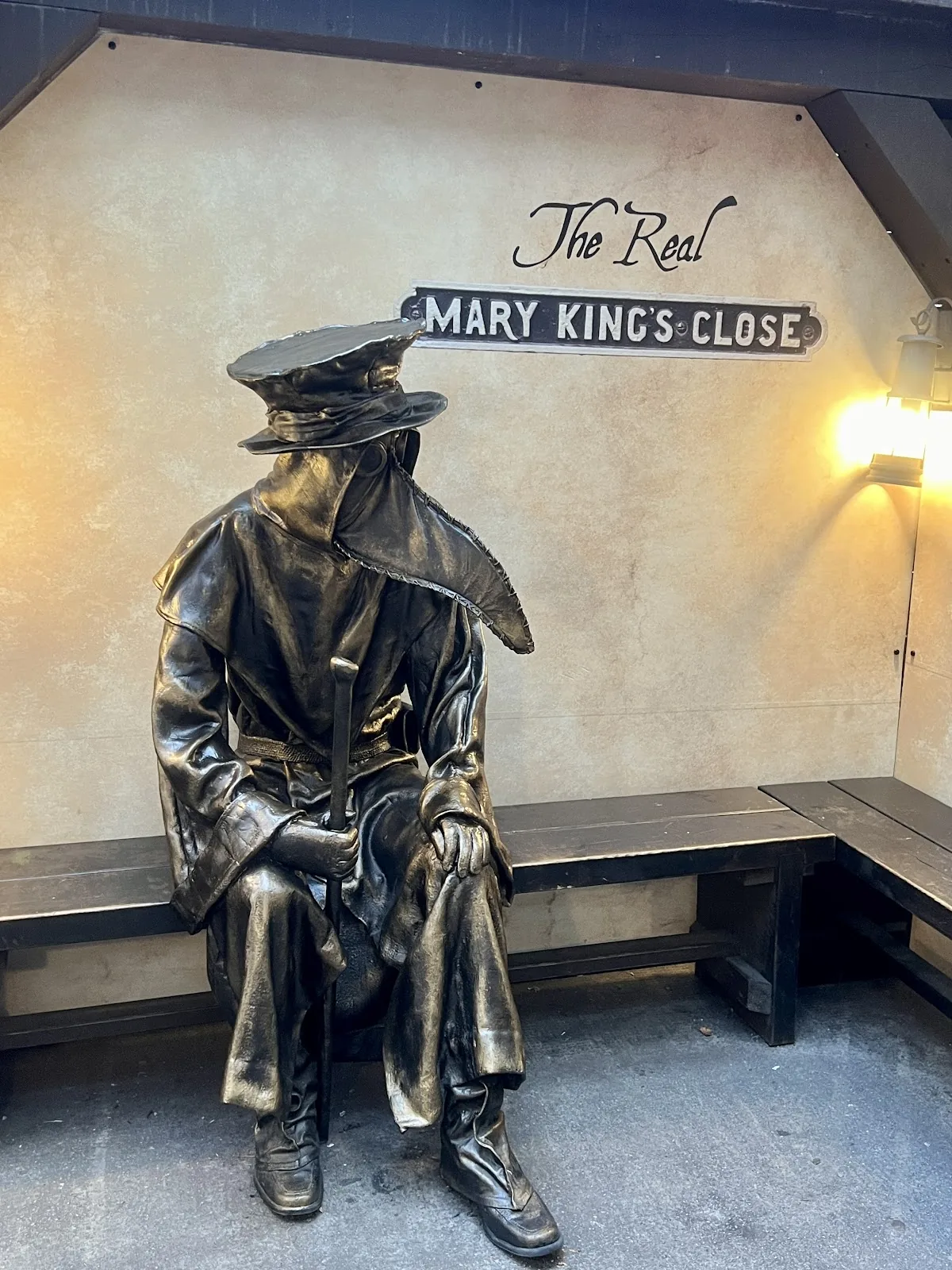 A bronze statue of a plague doctor in a beaked mask and wide hat, sitting on a bench under the Mary King’s Close sign