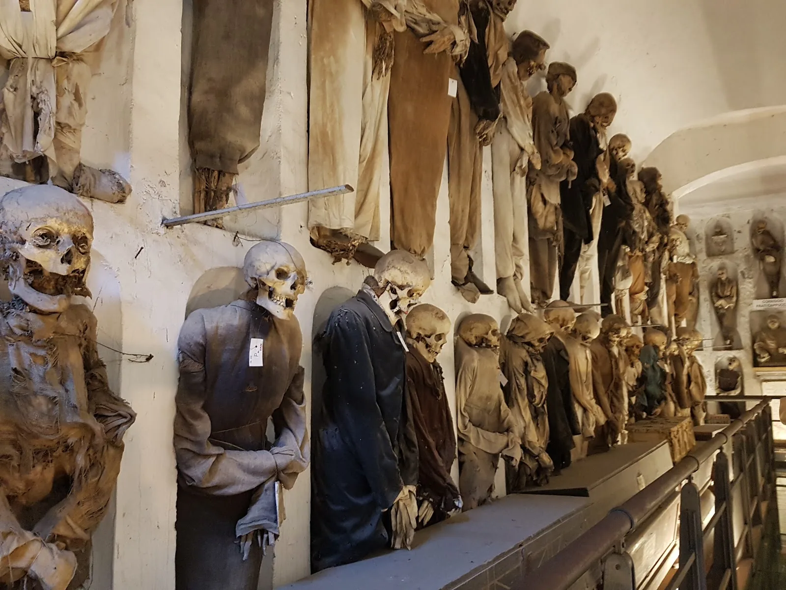 A long row of mummified bodies in clothes and robes standing in niches along a white corridor of the Capuchin Catacombs in Palermo