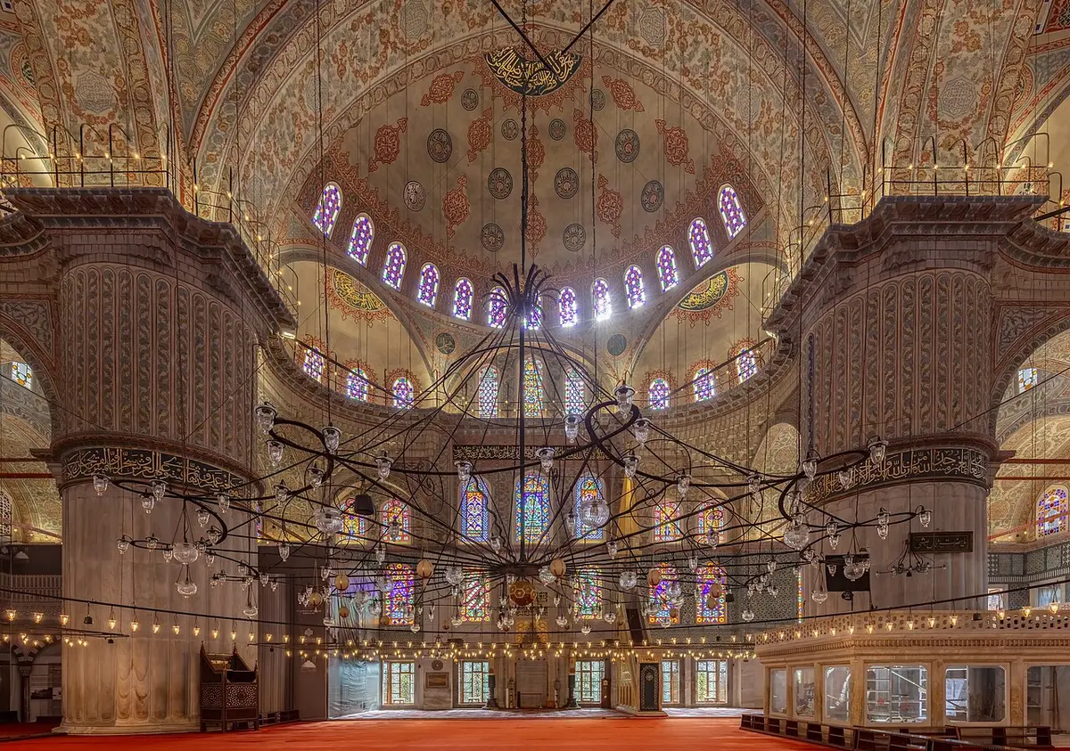 The interior of the Blue Mosque: domes and arches covered in painted patterns above a low ring of lamps and stained glass windows
