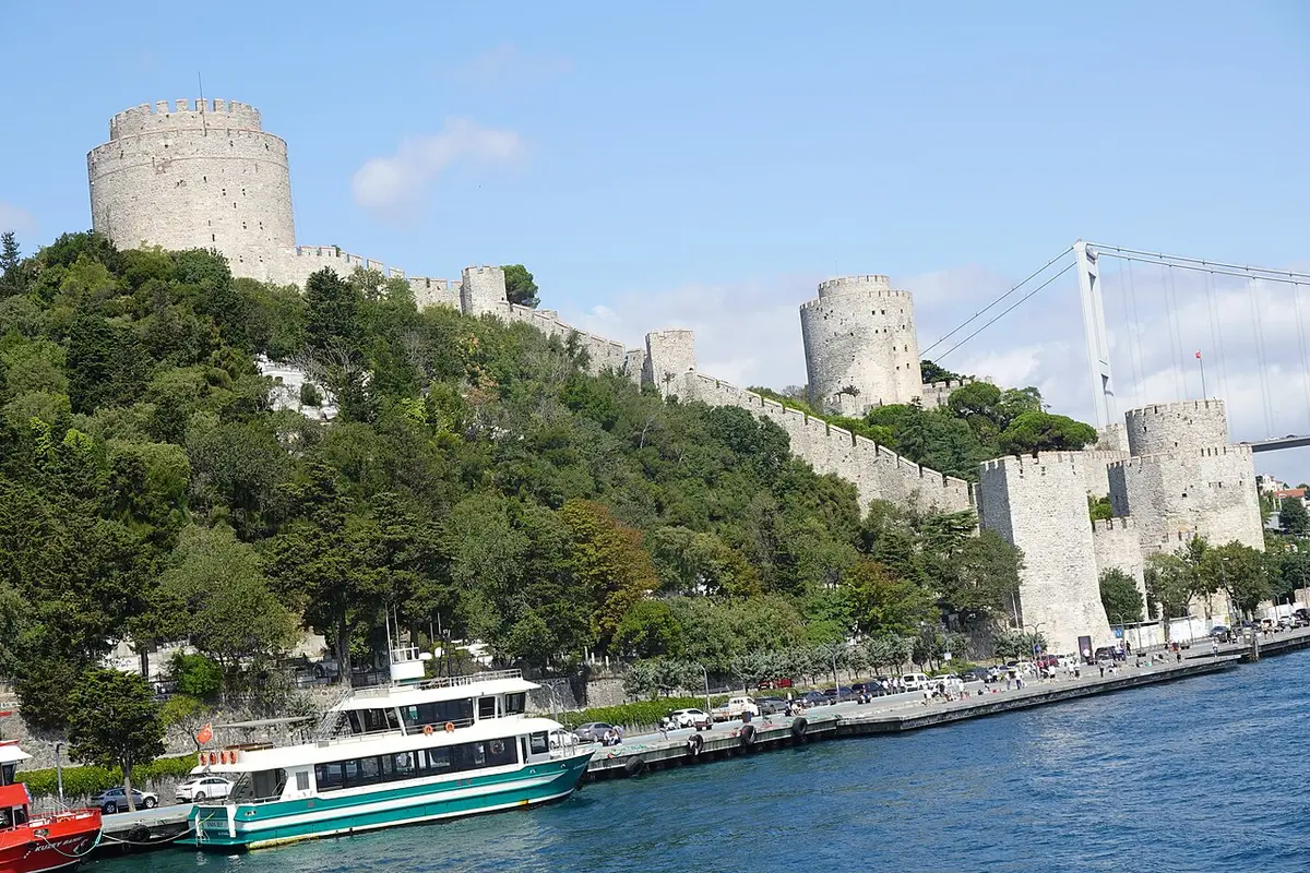 The towers and walls of Rumeli Hisarı climbing a wooded slope above the Bosphorus, a ferry at the quay and a suspension bridge behind
