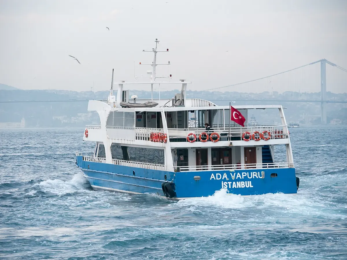A blue and white island ferry crossing choppy water, a suspension bridge in the haze behind