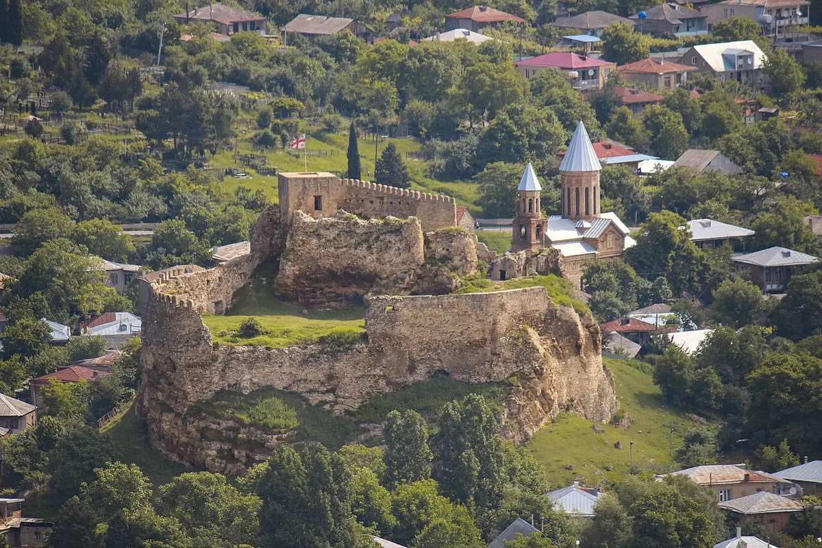 The ruined walls of Surami Fortress on a rocky outcrop among trees, a church with a conical dome beside it