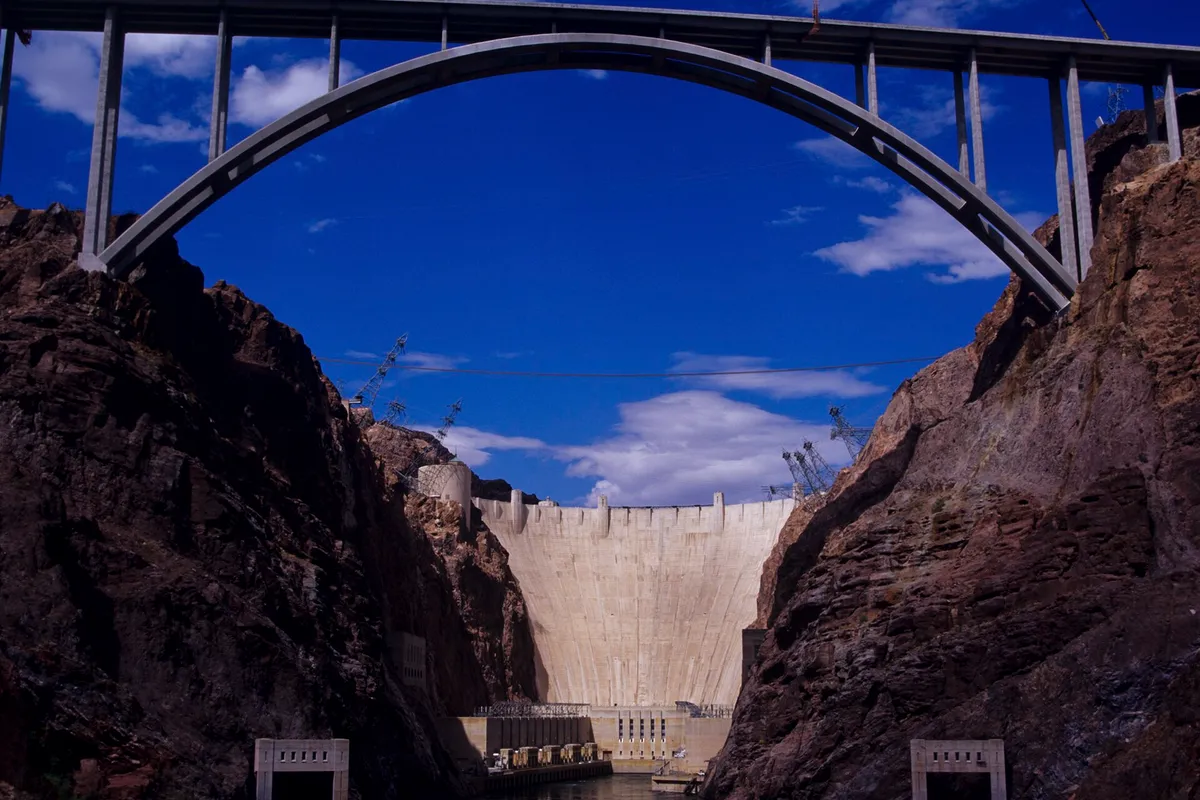 The concrete arch of a high bridge spanning a rocky canyon, Hoover Dam in the gap below