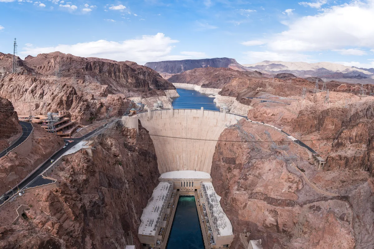 Hoover Dam from high above, its curved concrete face between canyon walls, the river below and Lake Mead beyond