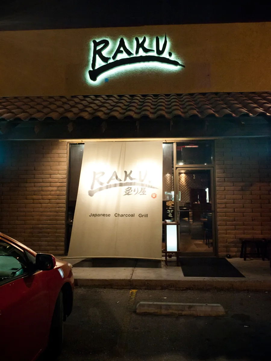 The lit Raku sign above a storefront at night, a white curtain at the door and a car parked in front