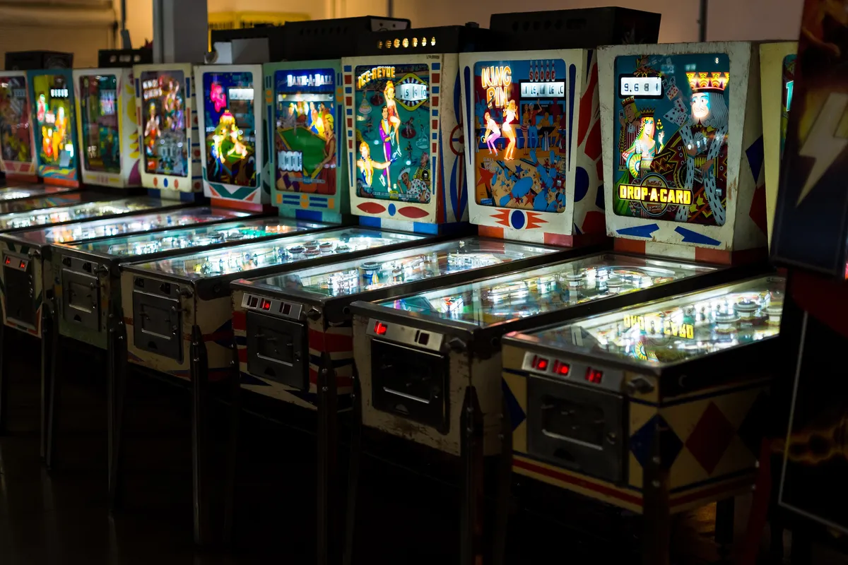 A long row of vintage pinball machines with lit backglasses in a dim hall