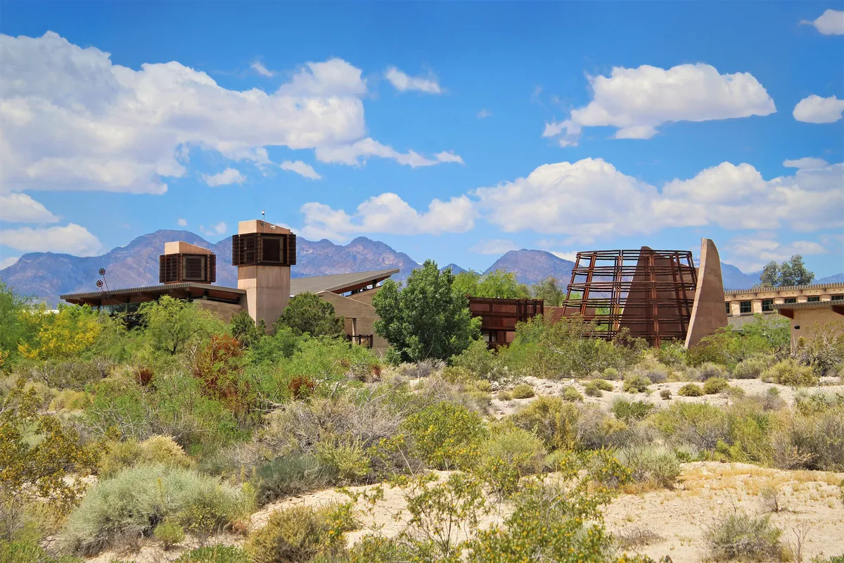 Low desert-colored buildings and rust-red towers among desert shrubs, mountains and white clouds beyond