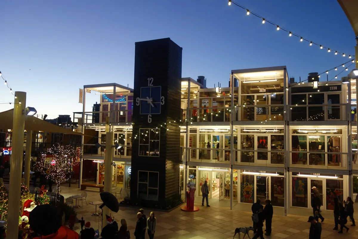 Stacked shipping-container shops with lit windows around a courtyard at dusk, strings of lights overhead