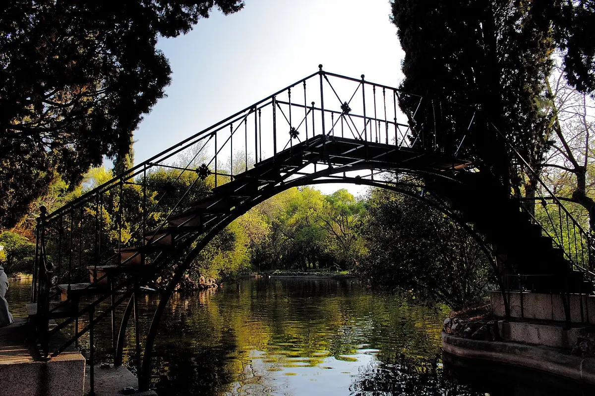 An iron footbridge arching over a pond among tall trees in El Capricho park