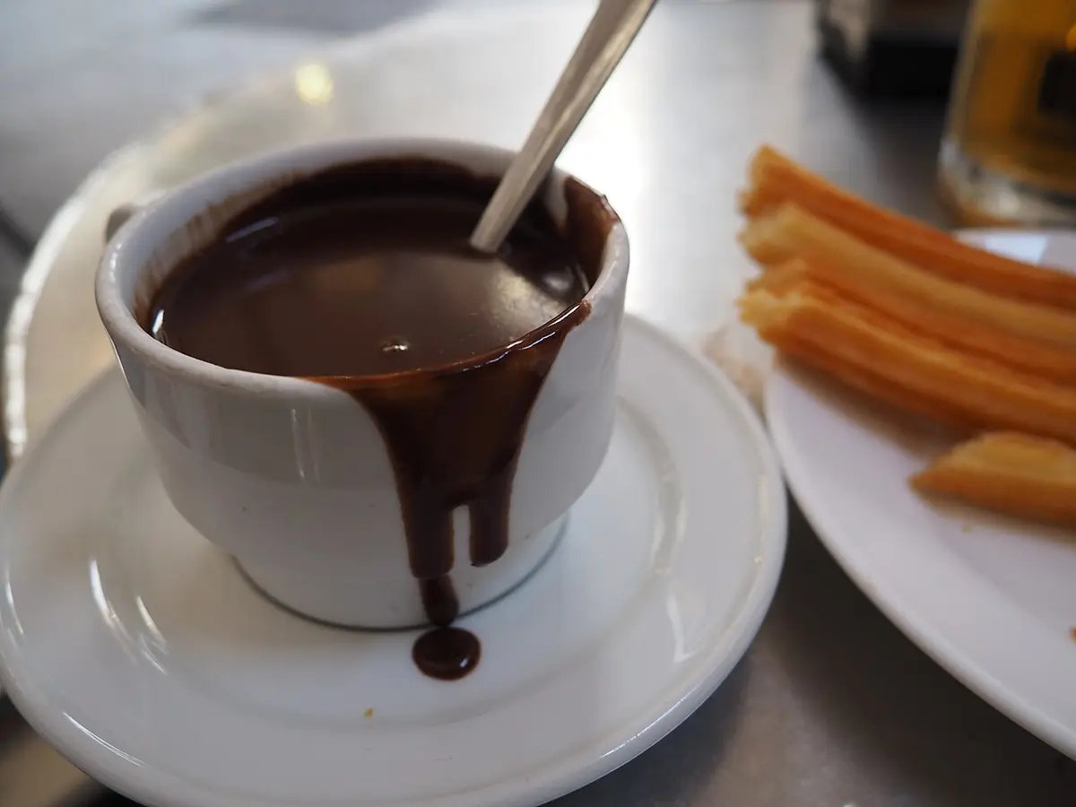 A cup of thick hot chocolate dripping down its side, with a plate of churros beside it