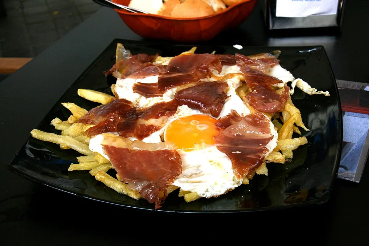 Fried eggs broken over fries and slices of cured ham on a black plate