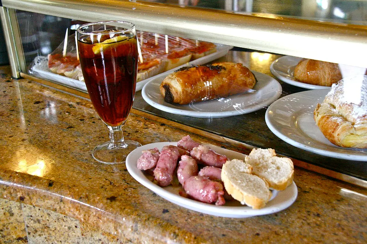 A glass of vermouth next to a small plate of sausages and bread on a bar counter