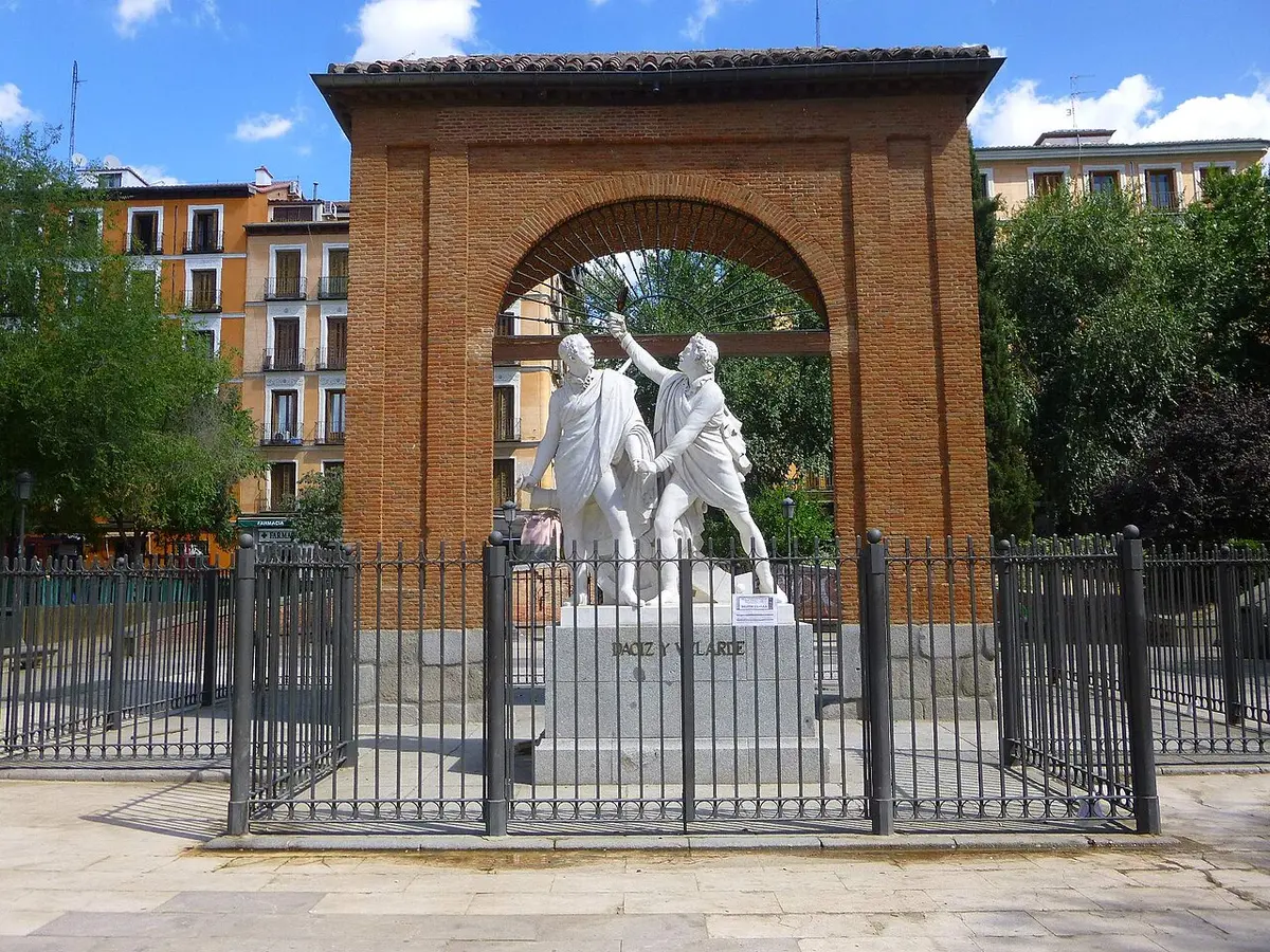 A brick arch in a square in Malasaña framing a white marble statue of two officers