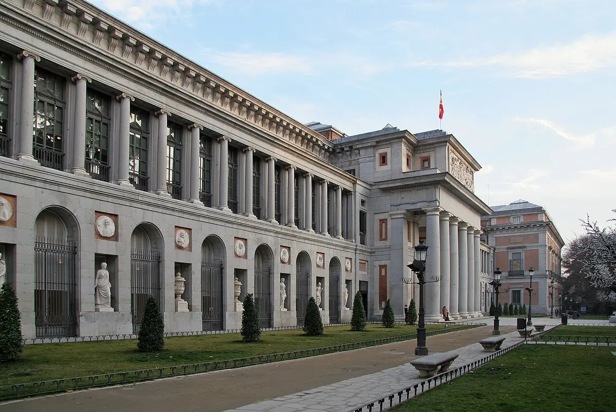 The long neoclassical west front of the Prado Museum, with its colonnade, behind a lawn and clipped hedges