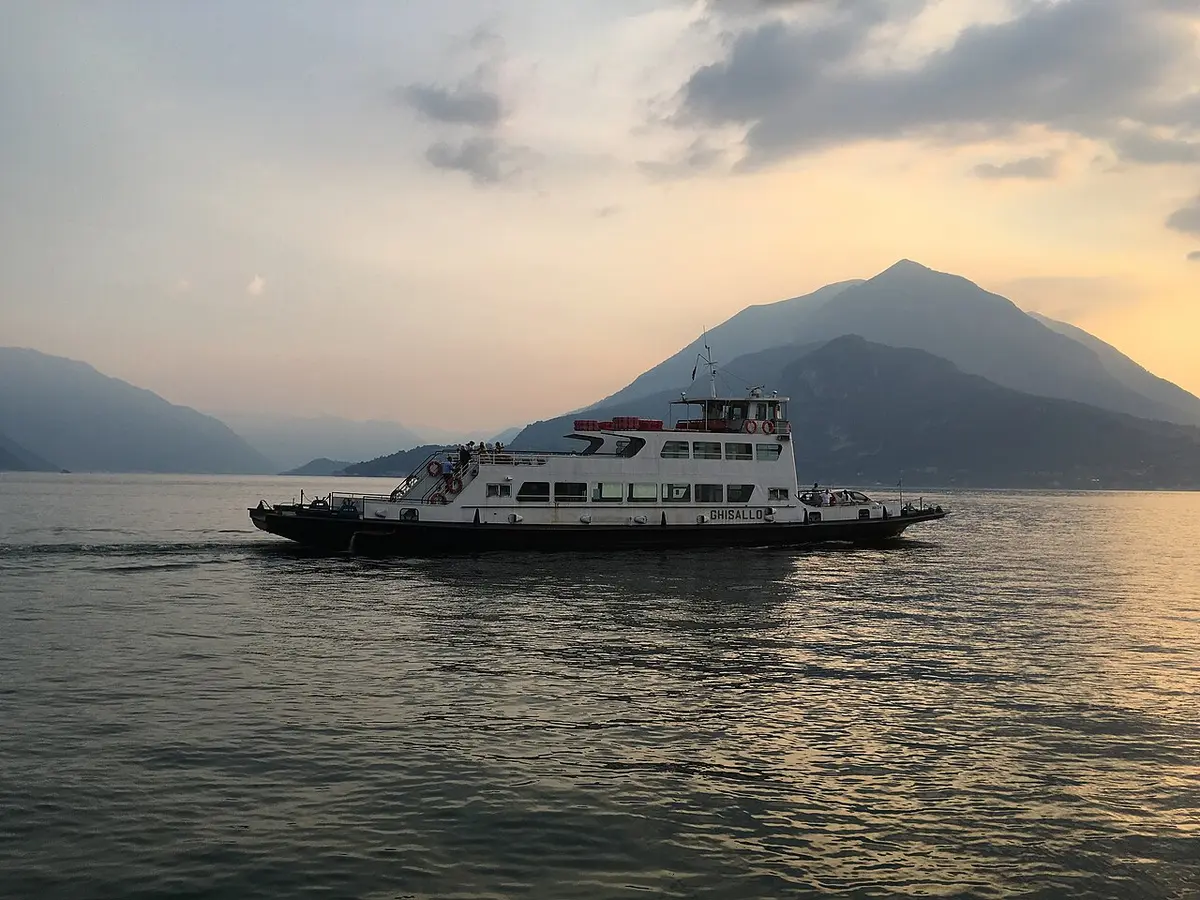 A white car ferry crossing Lake Como at dusk, mountains behind