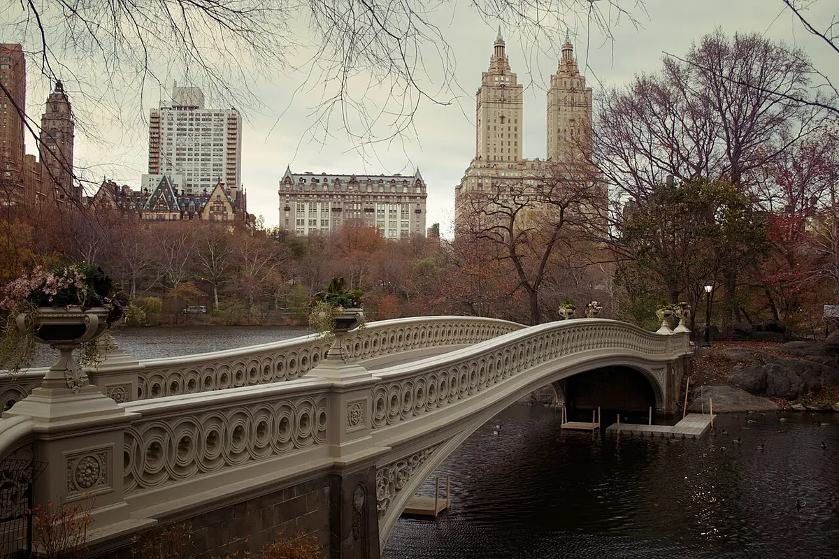 Bow Bridge crossing the Lake in Central Park in autumn, with the twin towers of the San Remo behind the trees