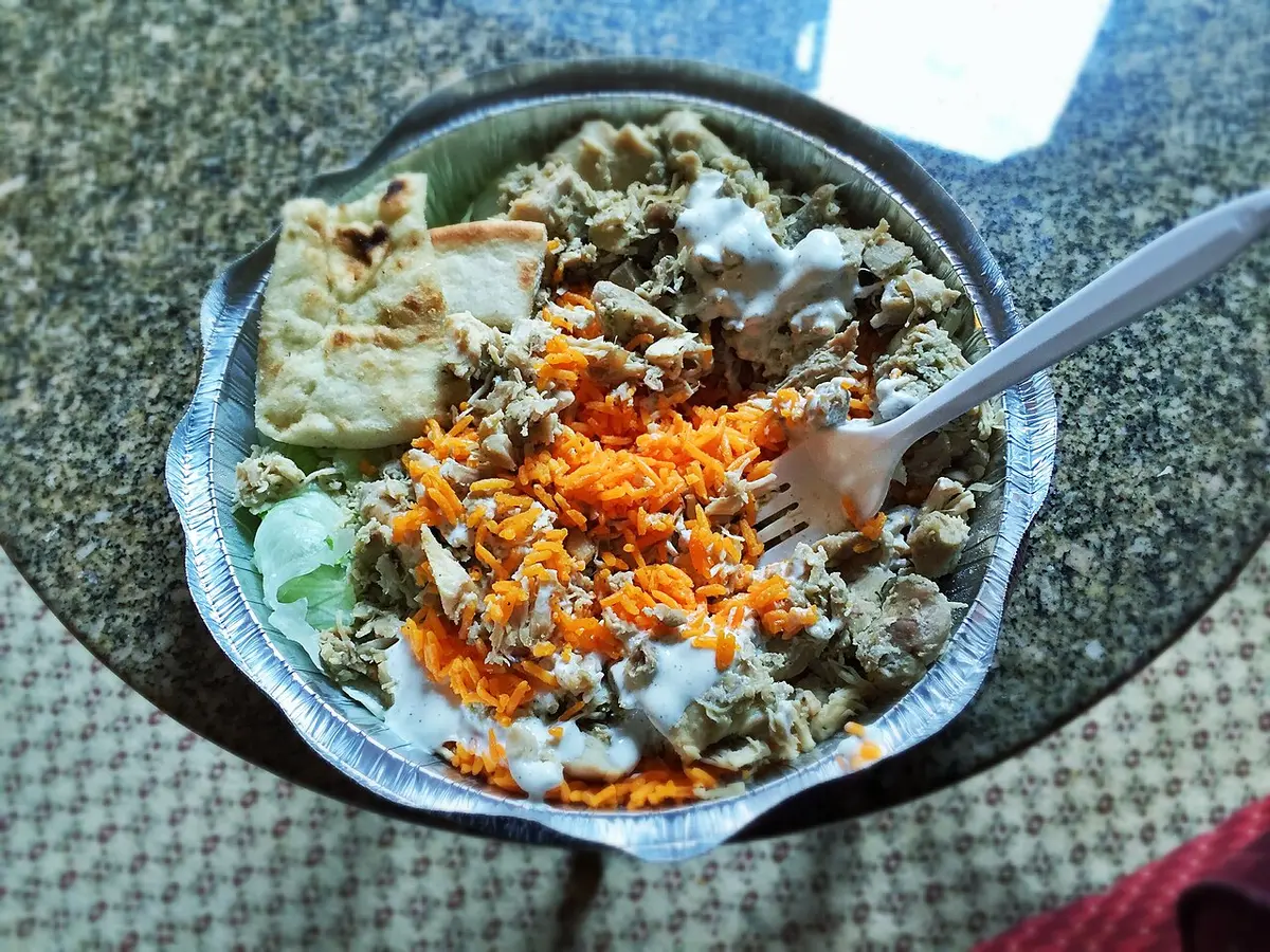 A foil tray of chicken over rice with lettuce, white sauce and pita