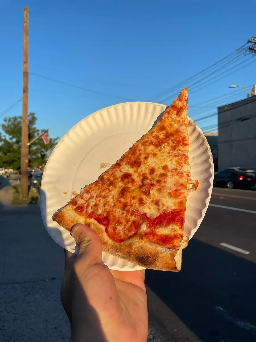 A slice of cheese pizza on a white paper plate, held up on a Queens street