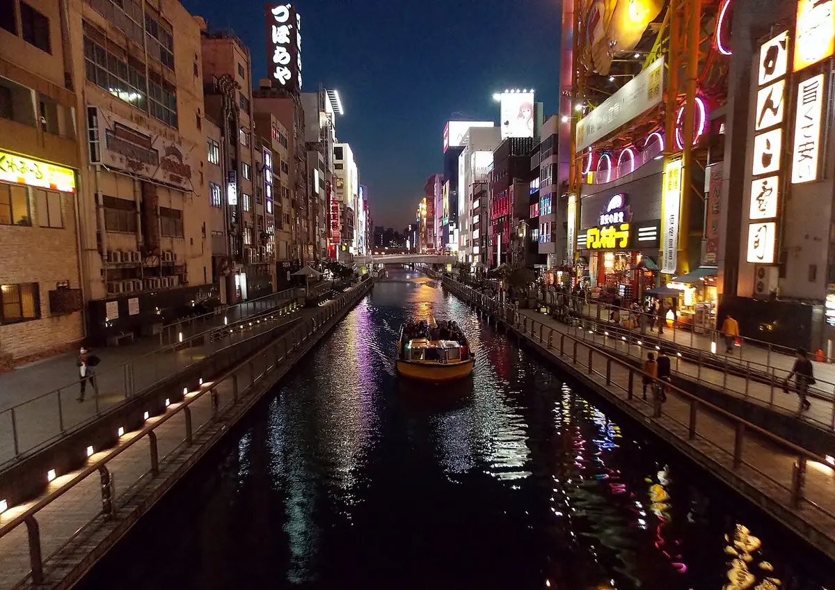 The Dōtonbori canal at dusk, lined with lit signs, a tour boat on the water
