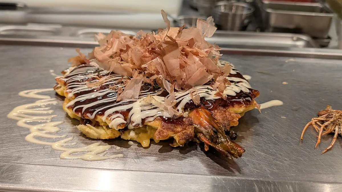 An okonomiyaki on a griddle, topped with sauce, mayonnaise and bonito flakes