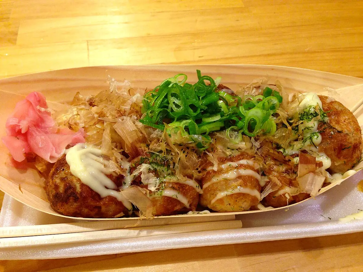 A paper boat of takoyaki topped with sauce, mayonnaise and green onion