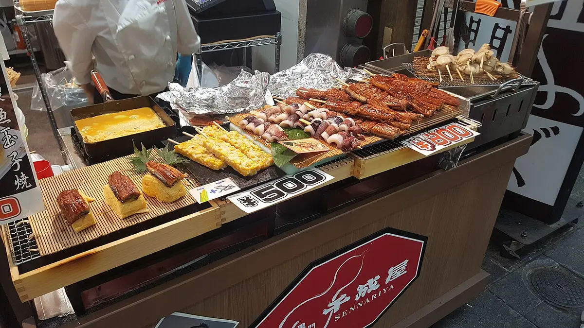 A stall at Kuromon Market with grilled seafood on skewers and trays of food for sale