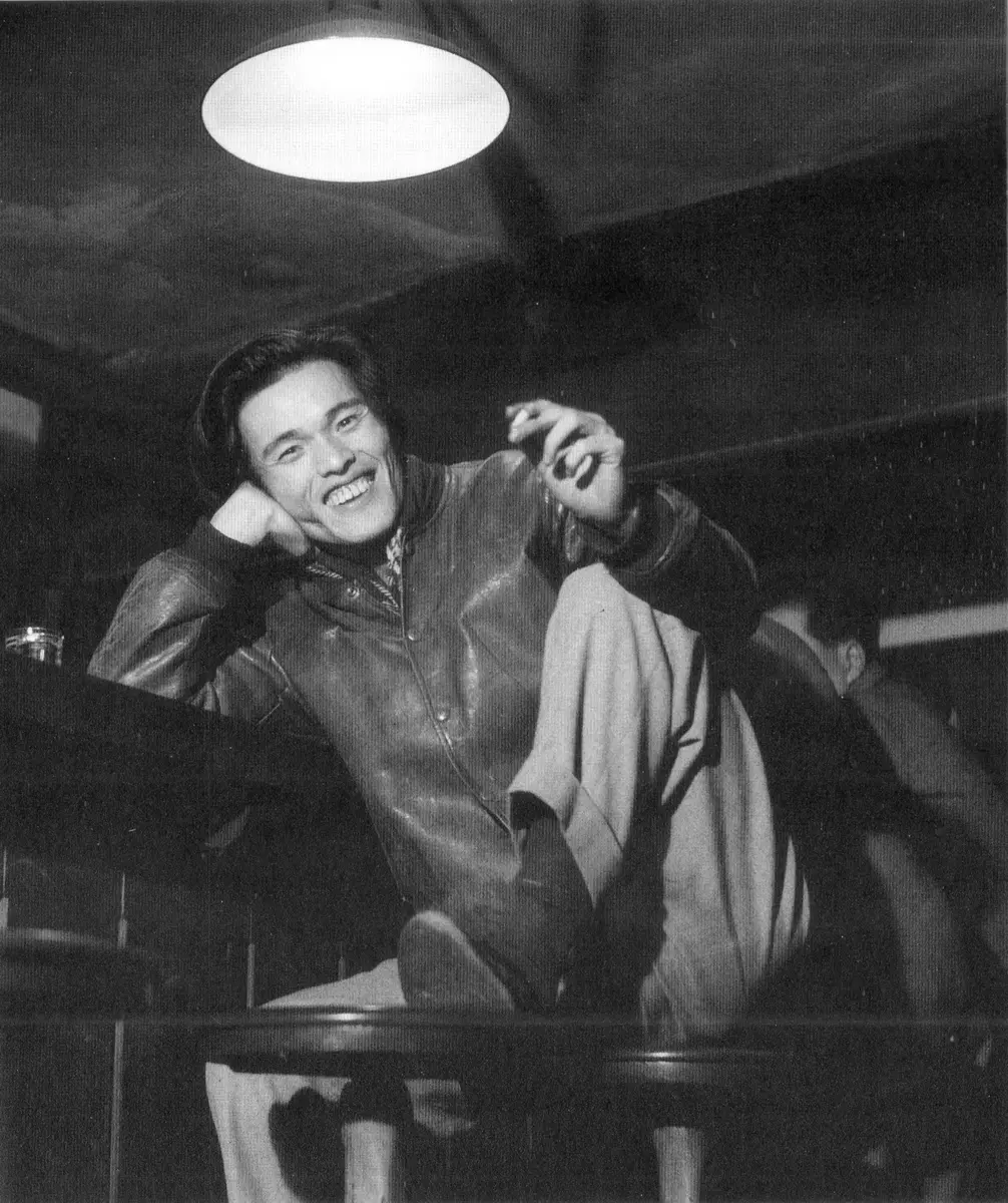 Black and white photograph of Oda Sakunosuke laughing at a bar counter, one knee raised